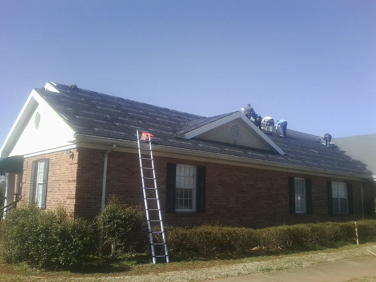 Crew tearing shingles off a brick ranch roof with a ladder set — Metal Roof Repair work by our crew in Mayville