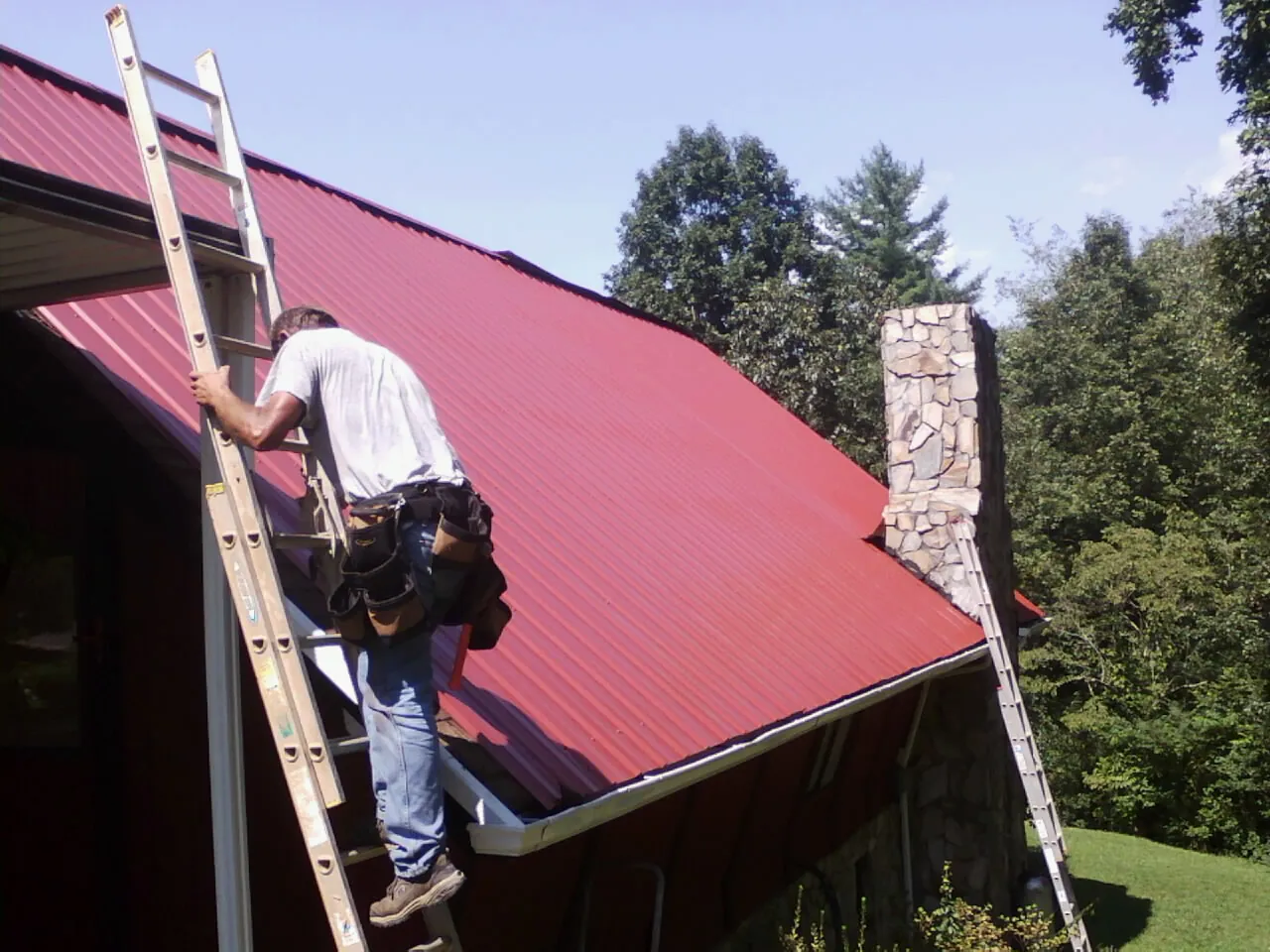 Roofer on a ladder beside a red metal roof and stone chimney — Soffit & Fascia Repair by Roofing Brothers in Mayville